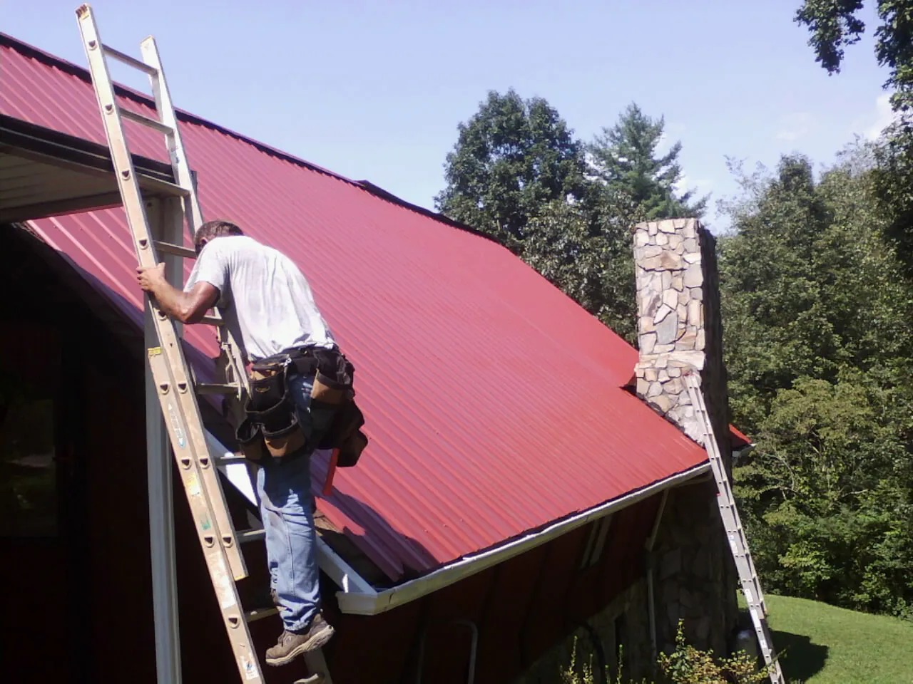 Roofer on a ladder beside a red metal roof and stone chimney — Soffit & Fascia Repair by Roofing Brothers in Klingerstown